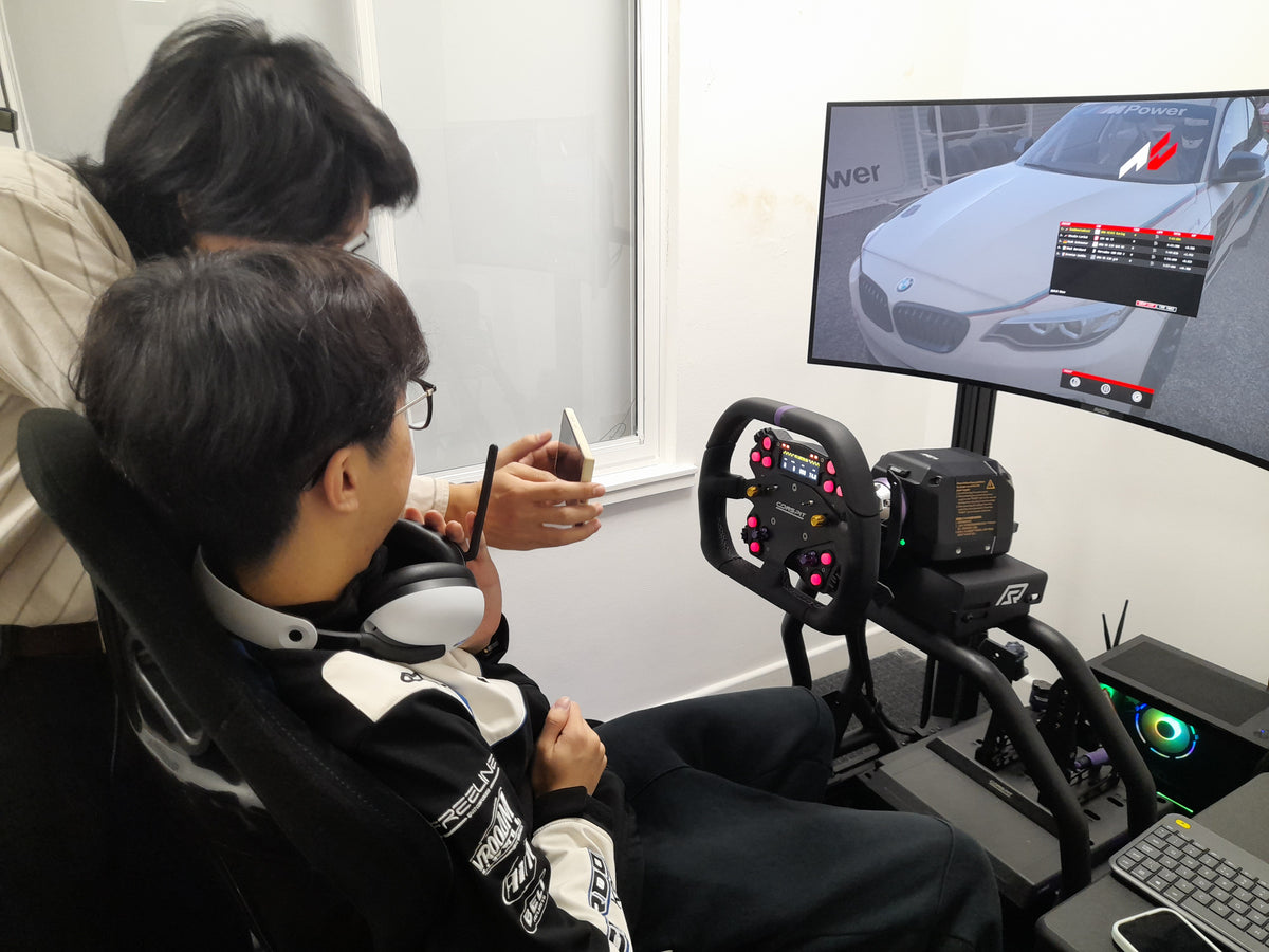 A person seated in a racing-style chair wearing over-ear headphones and a black-and-white racing jacket, using a steering wheel simulator attached to a curved monitor showing a car racing game. Another person stands behind holding a phone.