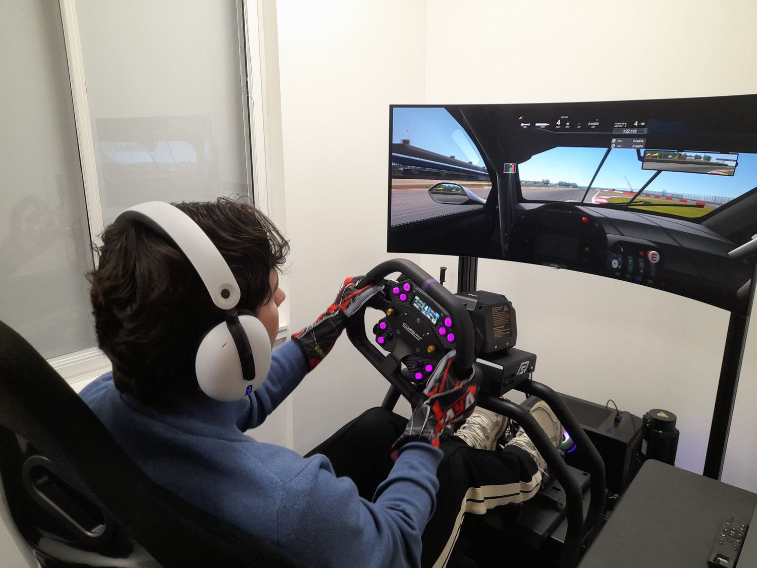 Person using a racing simulator with a steering wheel and headphones, facing a large screen displaying a racing game.